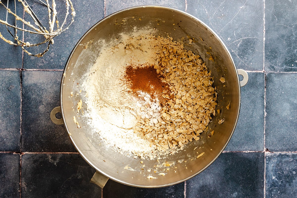 A mixing bowl containing flour, oats, cinnamon, and baking soda on a dark tiled countertop.