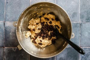 A metal mixing bowl with oatmeal cookie dough and raisins, with a black spatula resting inside, sits on a tiled countertop.