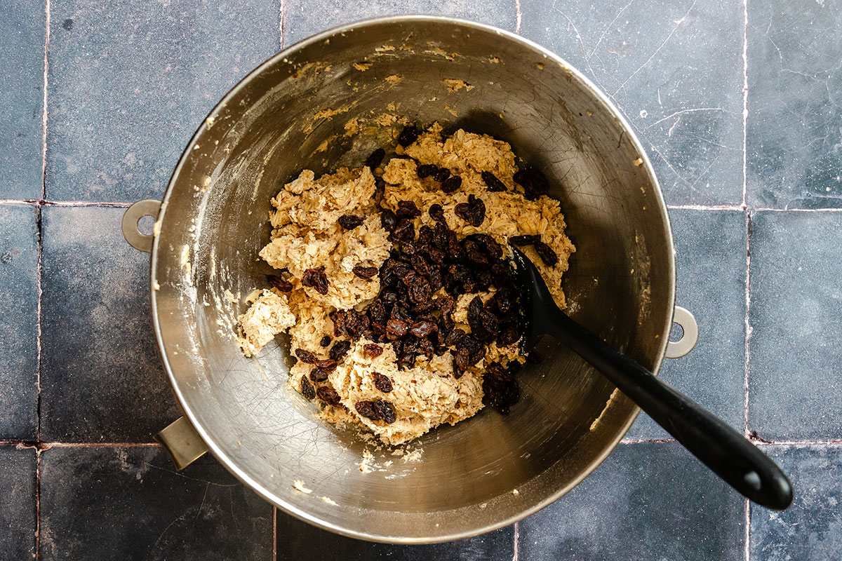 A metal mixing bowl with oatmeal cookie dough and raisins, with a black spatula resting inside, sits on a tiled countertop.
