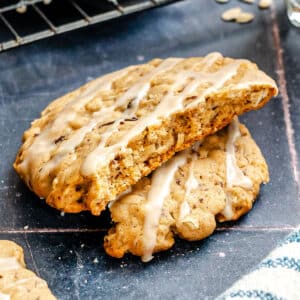 A large oatmeal cookie with icing drizzle, broken in half, is displayed on a dark surface with a cooling rack in the background.