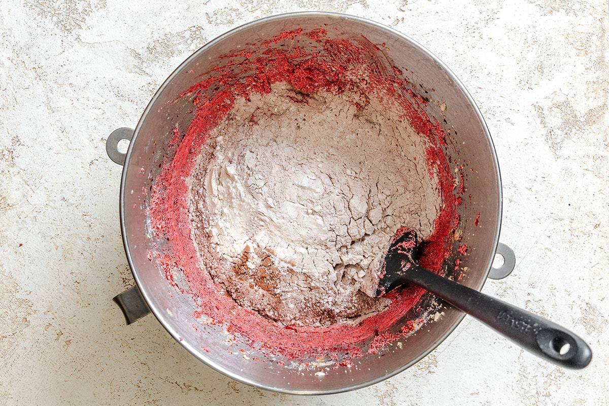 A metal mixing bowl containing flour and a red batter mixture, with a black spatula resting inside, sits on a light, textured surface.