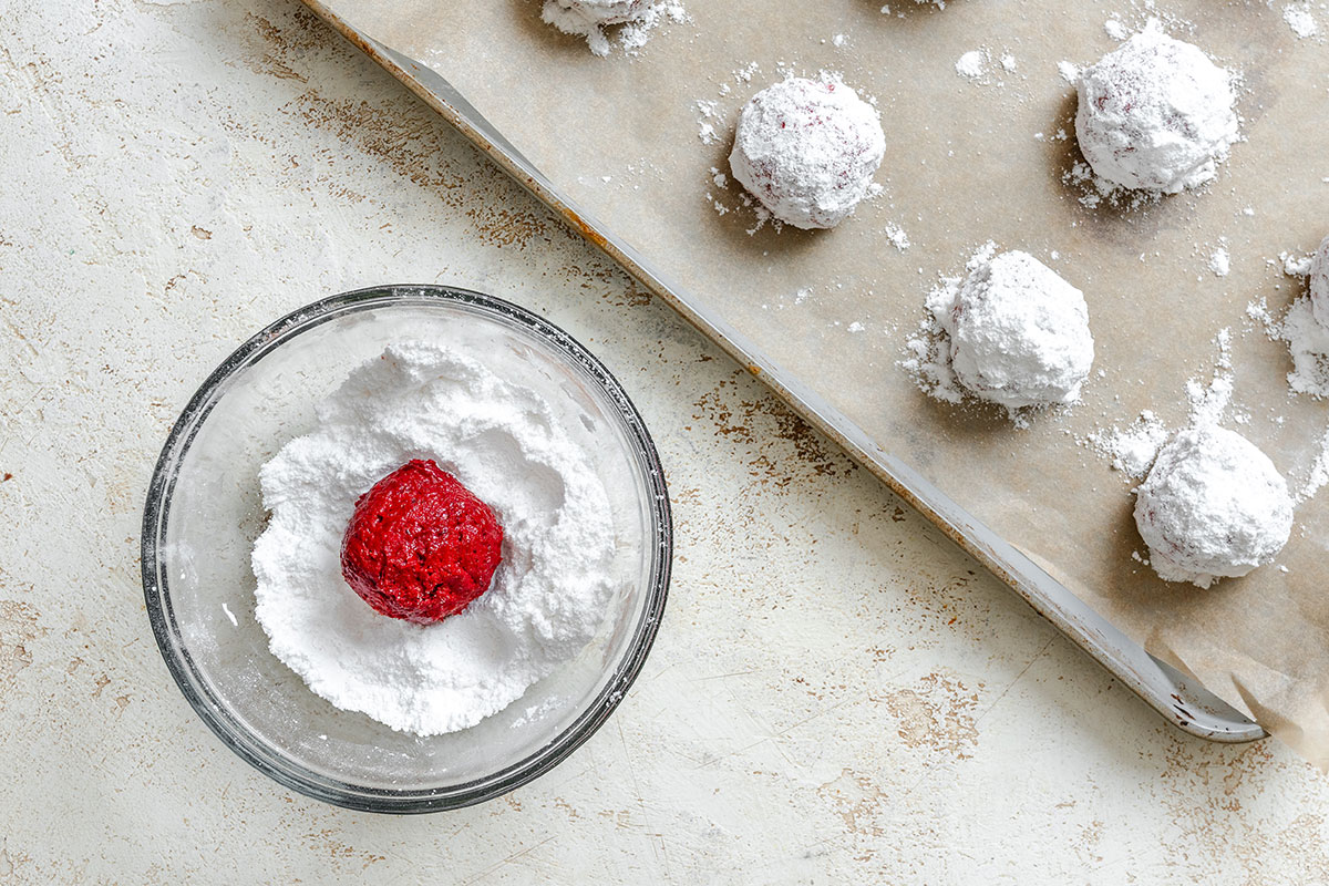 A bowl with a red dough ball coated in powdered sugar and a baking tray with several powdered sugar coated dough balls on parchment paper.