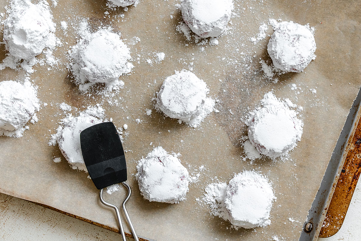 A baking tray lined with parchment paper holds several round cookies coated in powdered sugar, with a black spatula resting among them.