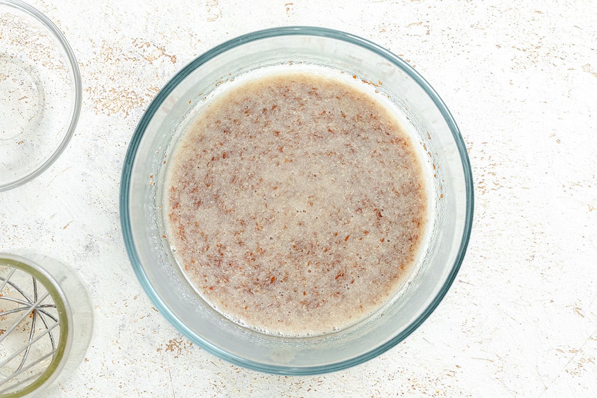 A glass bowl containing a mixture of ground flaxseed and water sits on a light-colored surface.