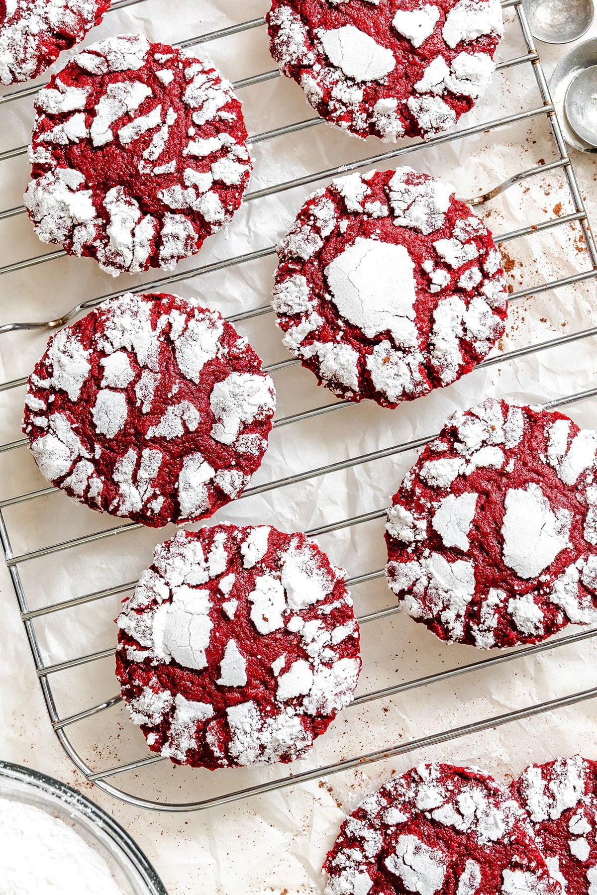 Red crinkle cookies covered with powdered sugar are cooling on a wire rack over a sheet of parchment paper.