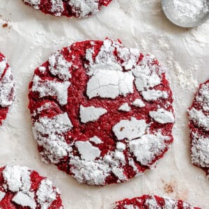 Red crinkle cookies with powdered sugar on top are arranged on parchment paper, with a metal scoop containing powdered sugar nearby.
