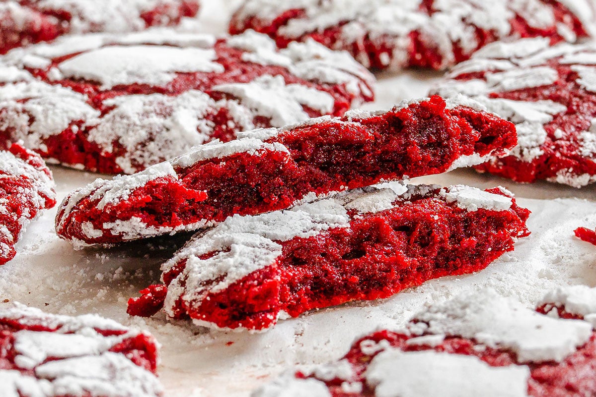 Red velvet crinkle cookies with a dusting of powdered sugar, one cookie broken in half to show its soft, red interior.