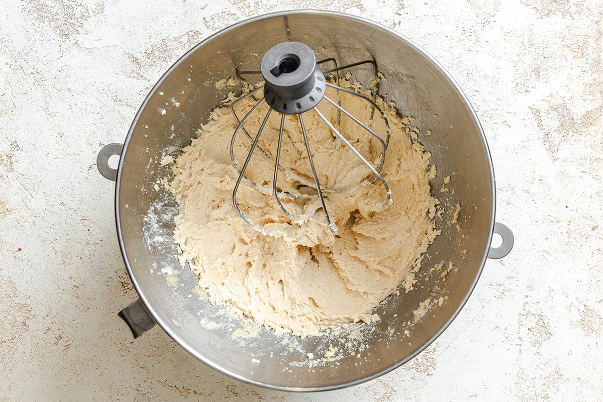 A metal mixing bowl with creamed dough and a whisk attachment sits on a light-colored surface.