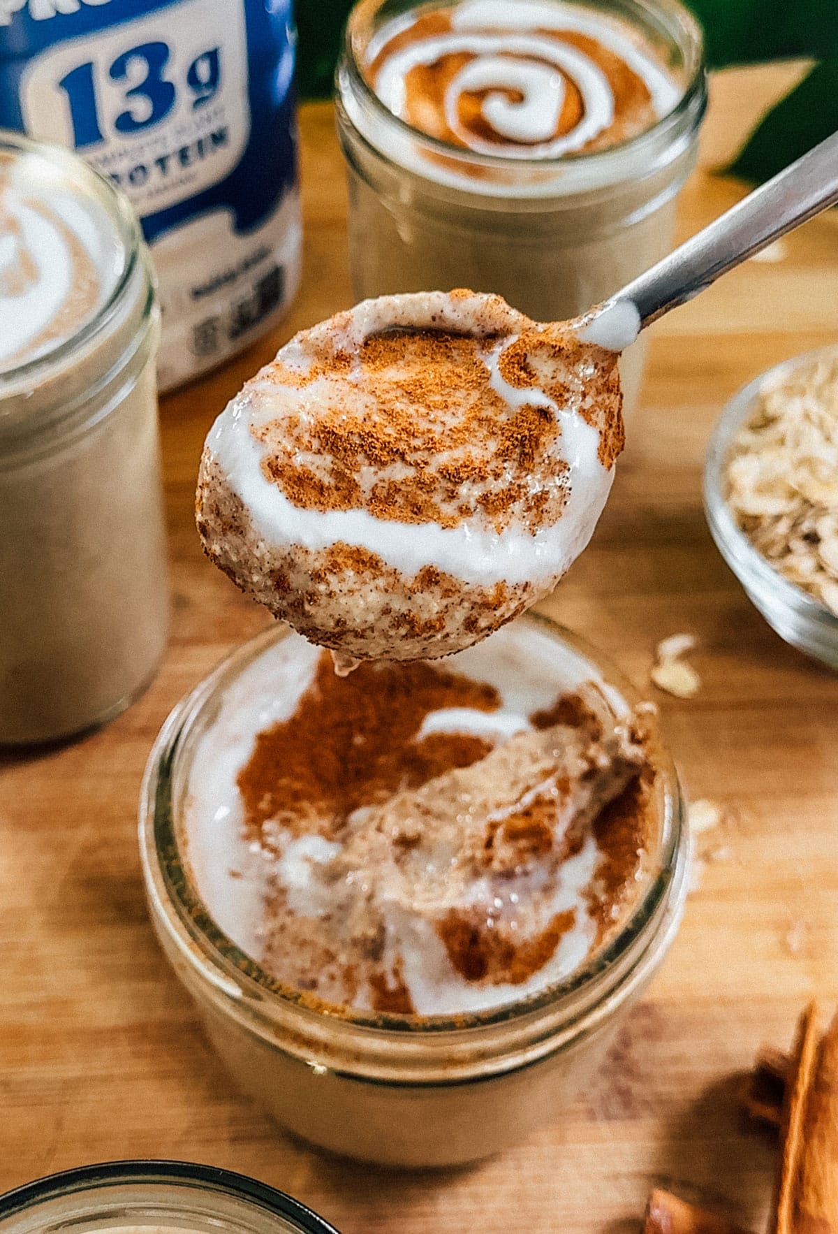 A spoonful of oatmeal topped with cinnamon and yogurt is held above a jar, surrounded by jars of similar oatmeal, rolled oats, and a container labeled 13g protein.