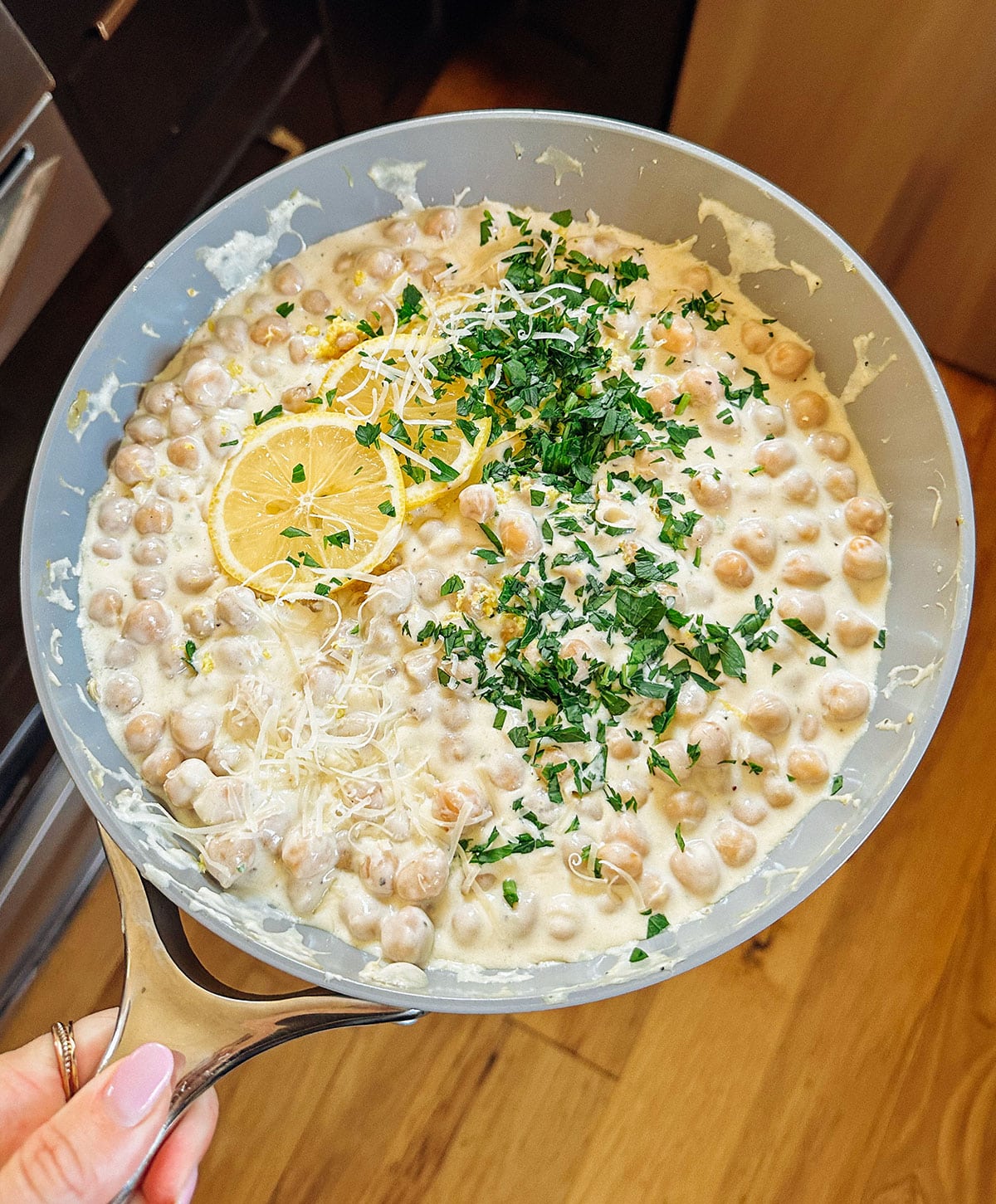 A skillet filled with creamy chickpeas, topped with grated cheese, chopped parsley, and lemon slices, held over a wooden floor.