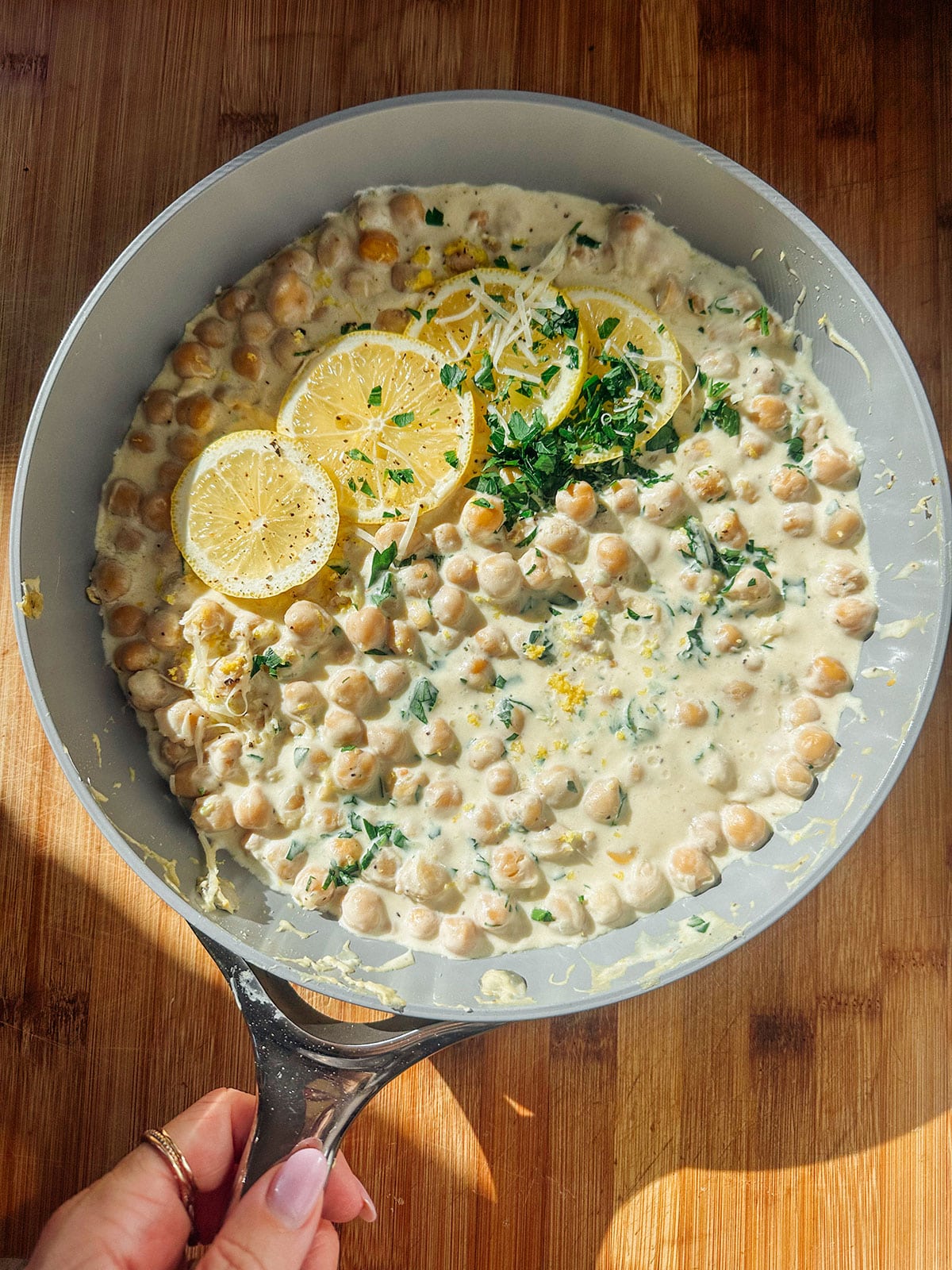 A skillet filled with creamy chickpeas, garnished with lemon slices, chopped herbs, and grated cheese, is being held over a wooden surface.