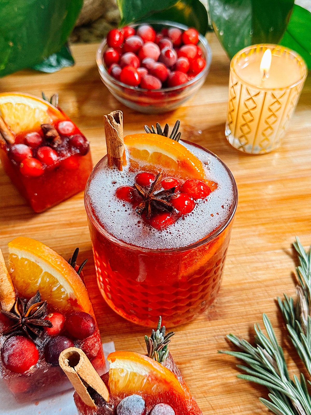 A glass of red cocktail garnished with orange slices, cranberries, cinnamon stick, and star anise, with a candle, cranberries, and rosemary on a wooden surface.