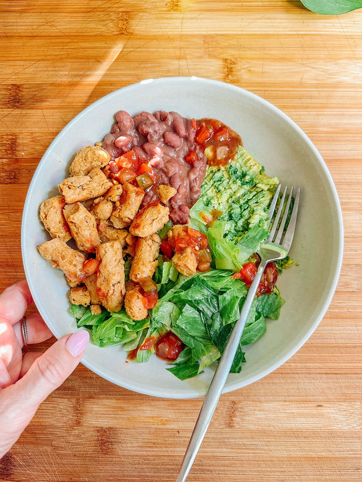 A bowl with seasoned chicken strips, pinto beans, guacamole, salsa, and chopped romaine lettuceโperfect for easy dinnersโwith a fork and a hand holding the bowl.