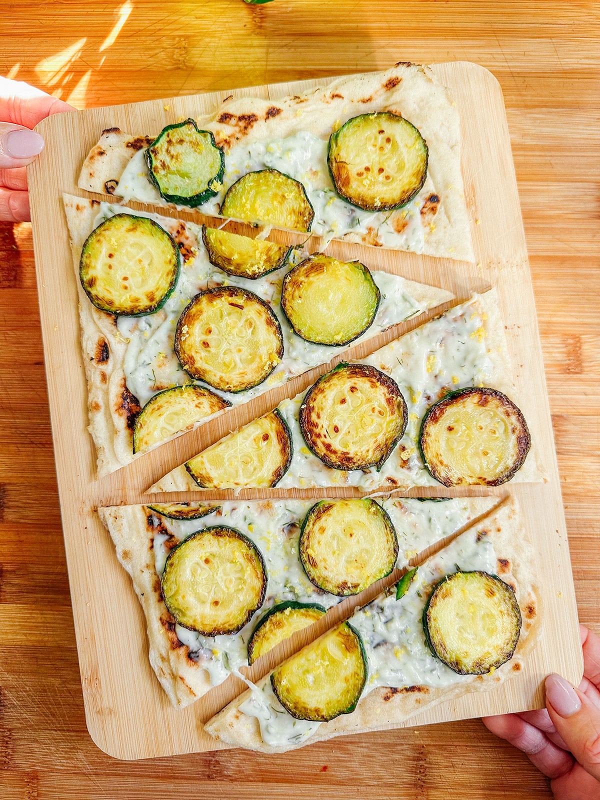Rectangular flatbread with white sauce and sliced zucchini, cut into six pieces, on a wooden board held by two hands over a wood surfaceโperfect for easy dinners or those seeking delicious vegetarian dinners.