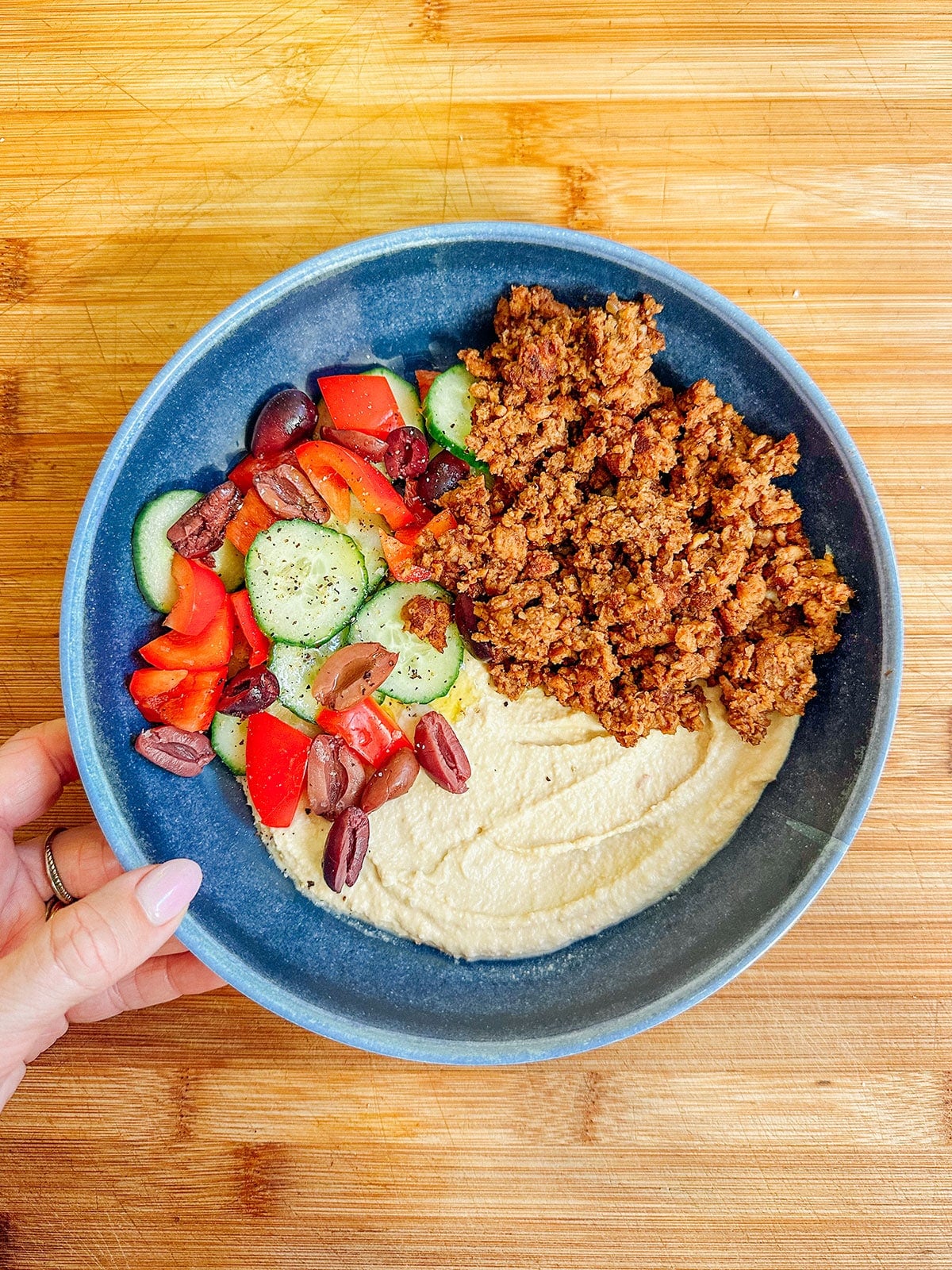 A blue bowl containing hummus, ground spiced meat, sliced cucumbers, tomatoes, and Kalamata olives on a wooden surface. A hand is holding the bowlโperfect for easy dinners or anyone looking for a quick meal after reading a Hungryroot review.