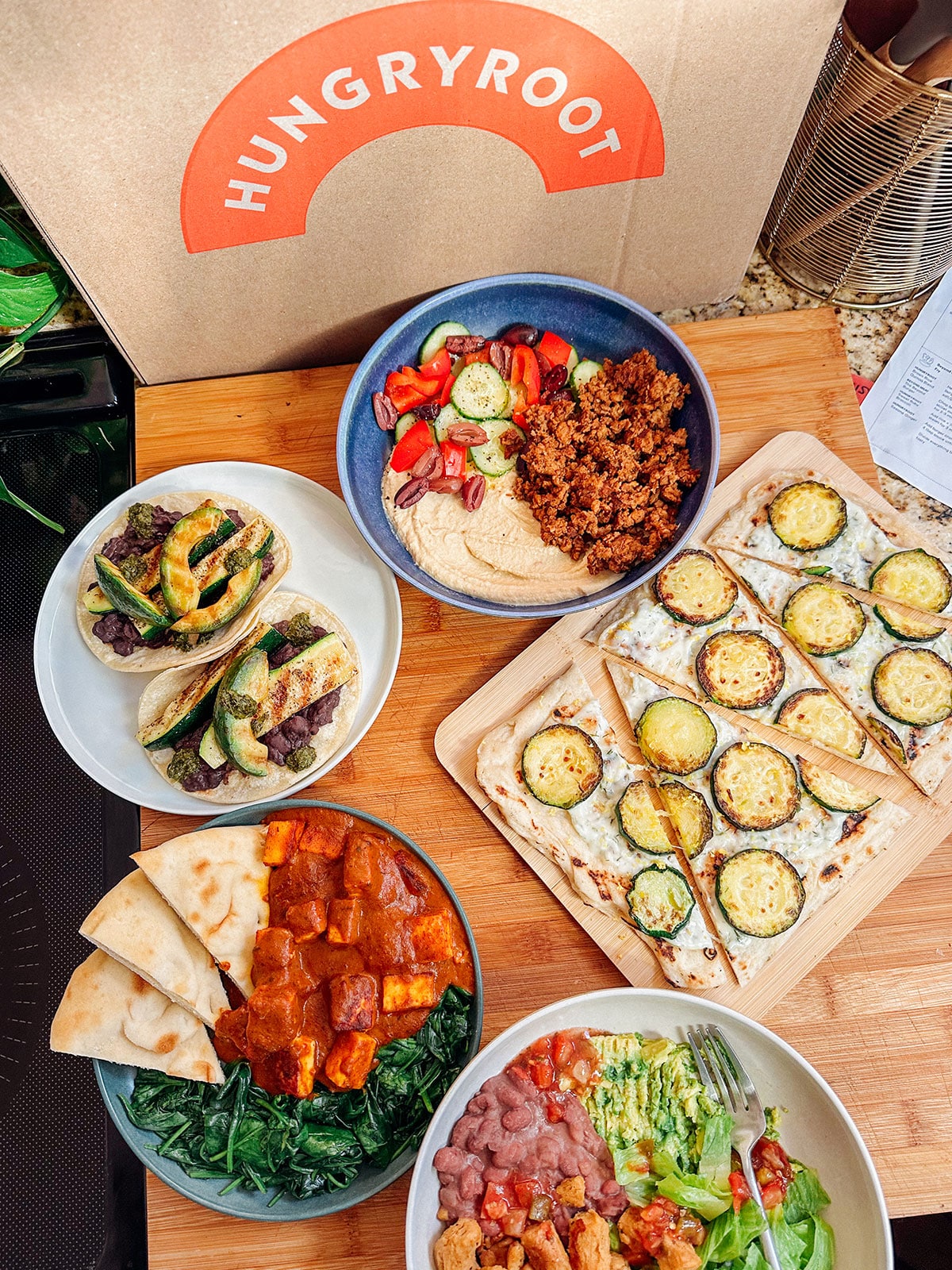 A variety of prepared mealsโlike tacos, a zucchini flatbread, a salad bowl, dip with pita, and greensโset on a wooden board next to a Hungryroot box, perfect for effortless and delicious vegetarian dinners.