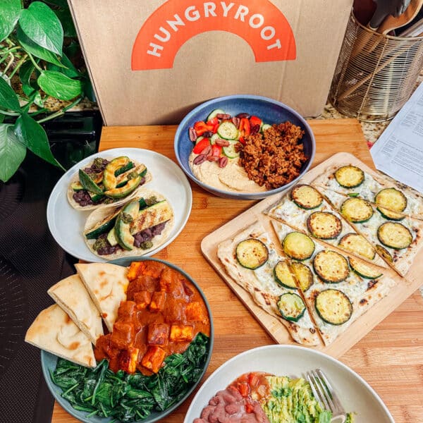 Various prepared dishesโlike tacos, flatbread with zucchini, salad, and a bowl with sauce and greensโare arranged on a wooden surface in front of a Hungryroot meal delivery box, showcasing easy dinners perfect for any Hungryroot review.