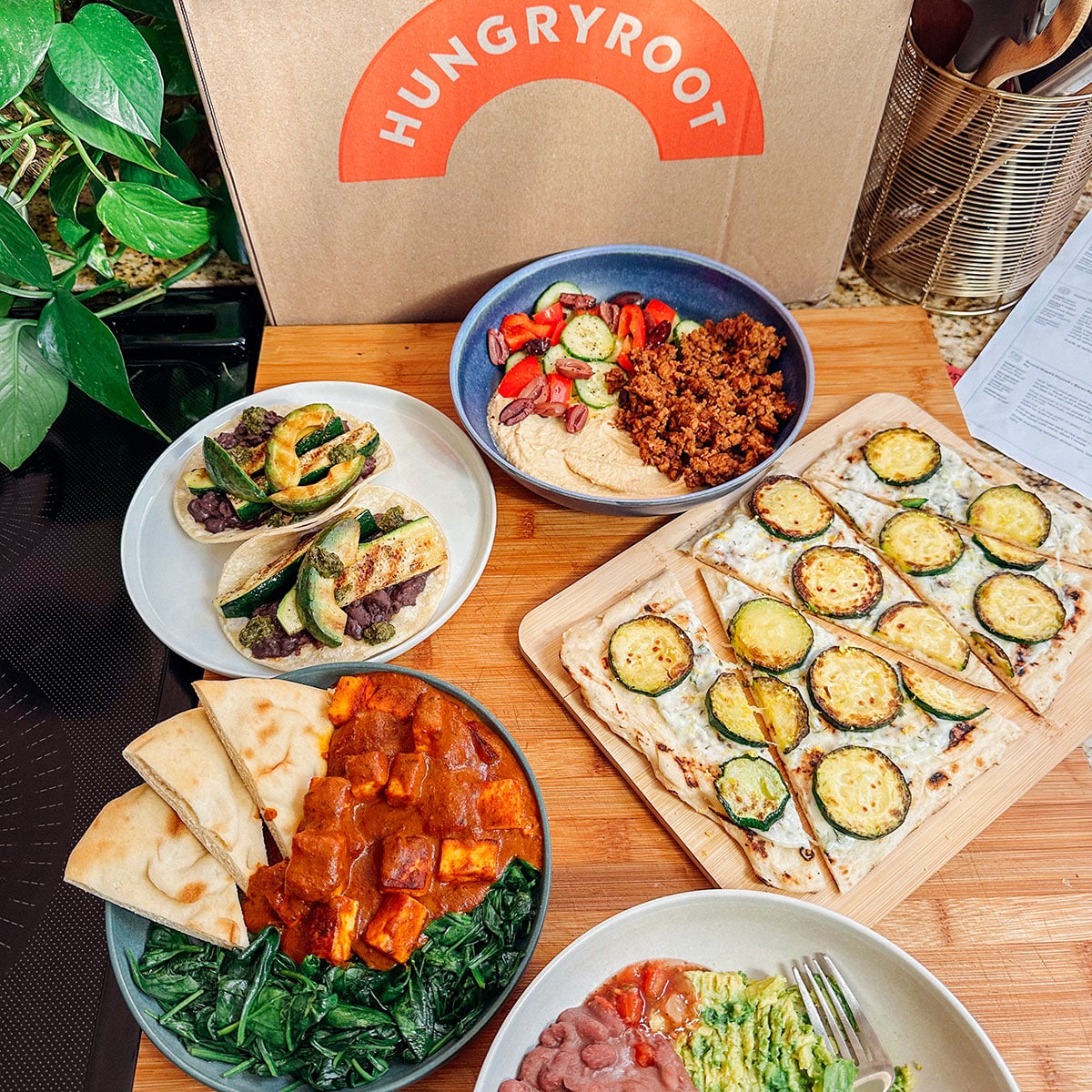 Various prepared dishesโlike tacos, flatbread with zucchini, salad, and a bowl with sauce and greensโare arranged on a wooden surface in front of a Hungryroot meal delivery box, showcasing easy dinners perfect for any Hungryroot review.