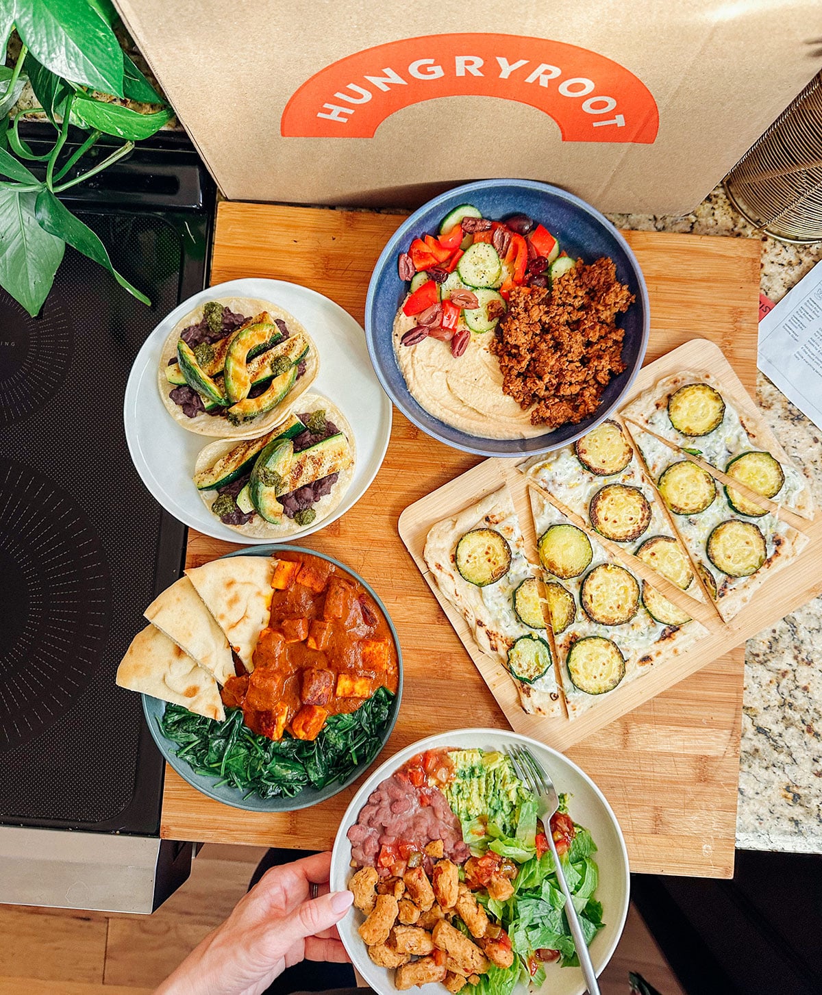 An overhead view of assorted plant-based meals in bowls and on a board, next to a Hungryroot delivery box on a kitchen counterโperfect inspiration for easy dinners or anyone seeking a thorough Hungryroot review.