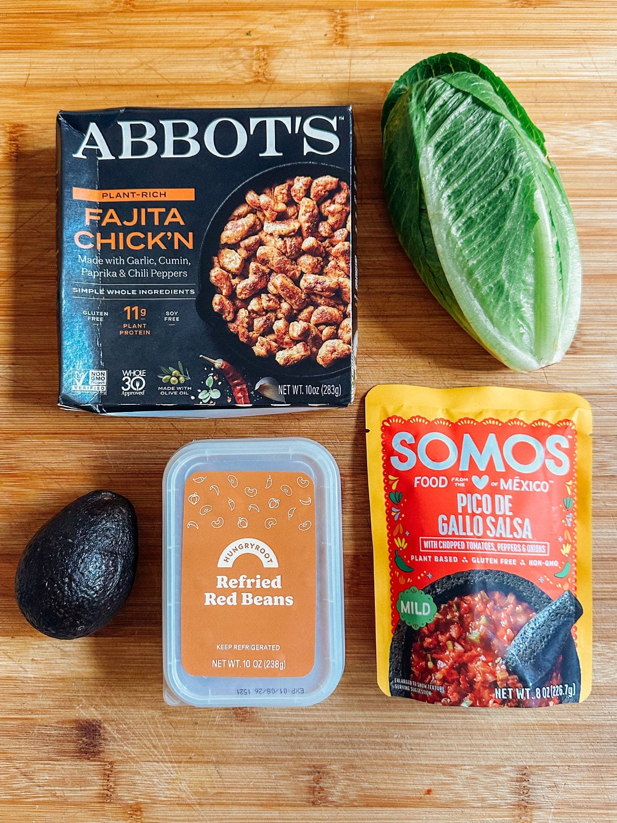 A package of plant-based fajita chicken, a pouch of pico de gallo salsa, refried red beans, half a romaine heart, and an avocado on a wooden surfaceโperfect for easy vegetarian dinners or your next Hungryroot review.
