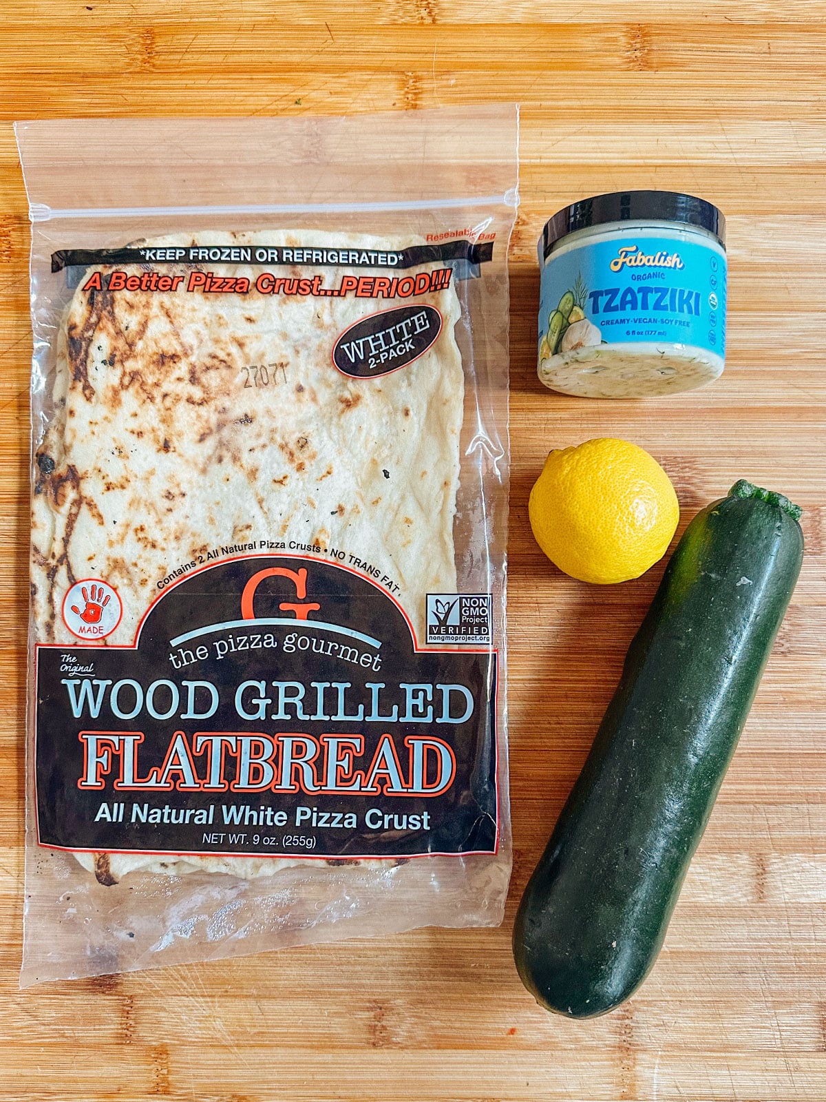 A package of wood grilled flatbread, a jar of tzatziki, a lemon, and a zucchini on a wooden surfaceโperfect for easy dinners and delicious vegetarian dinners.
