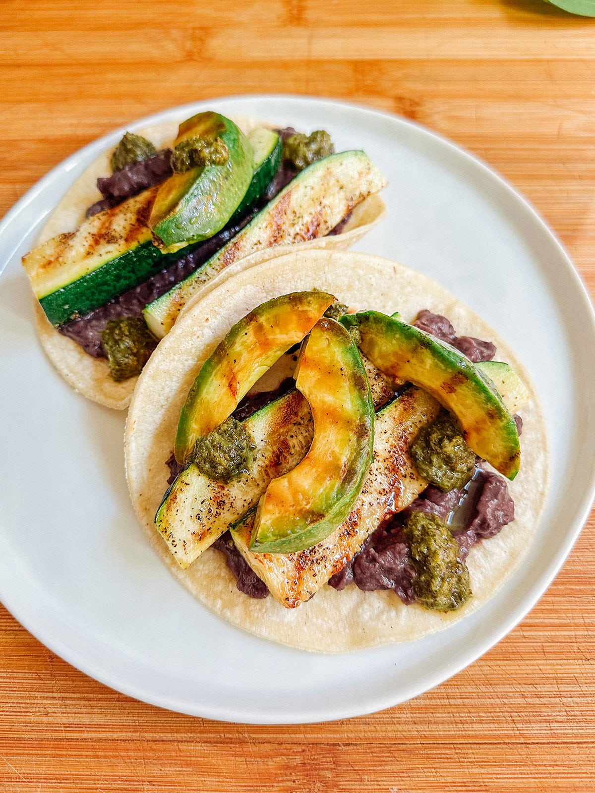 Two corn tortillas topped with black bean spread, grilled zucchini slices, and dollops of green sauce make for delicious vegetarian dinnersโperfect for easy dinners on busy nights. Served on a white plate set on a wooden surface.