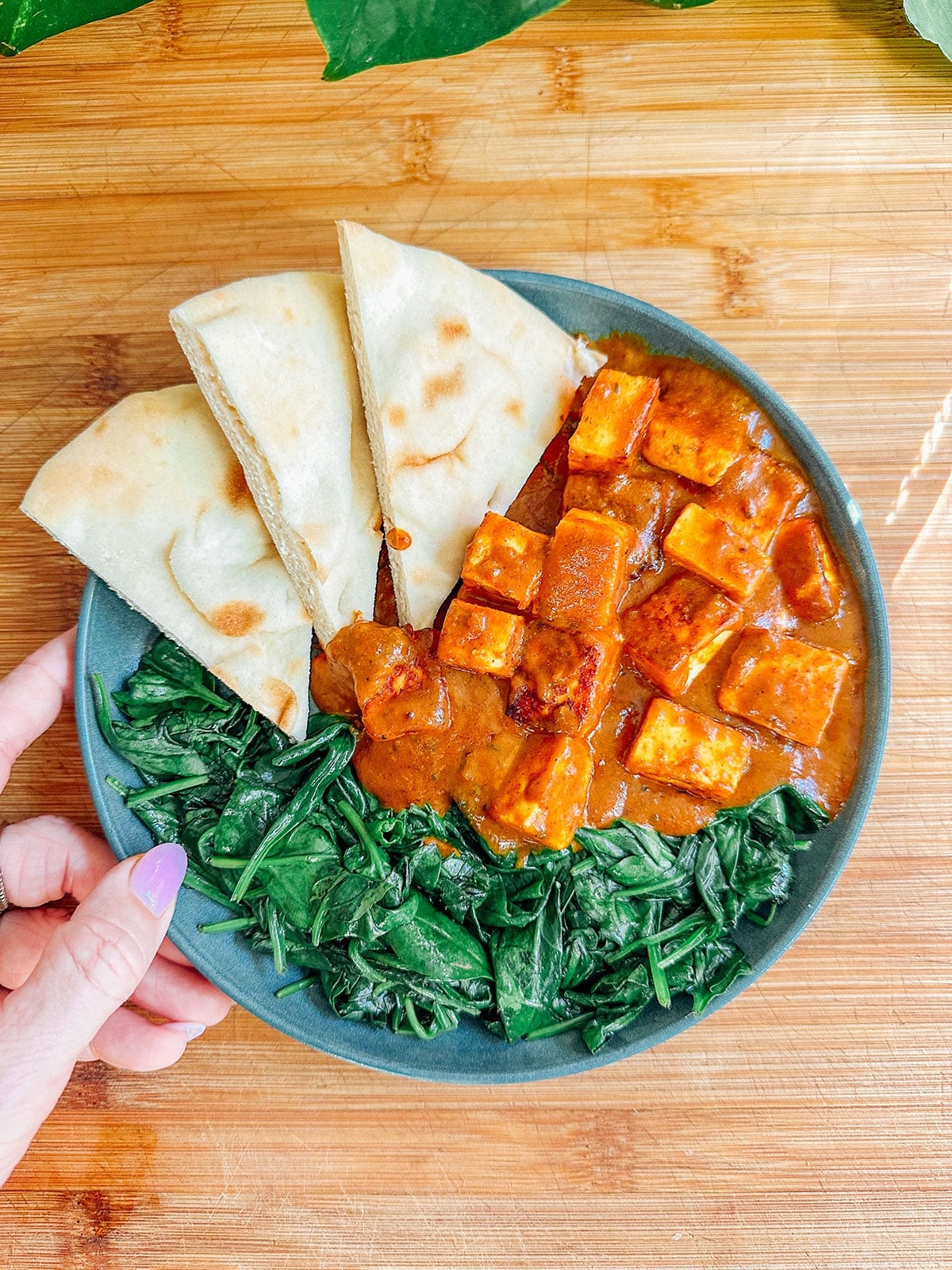 A plate with sautรฉed spinach, paneer in red curry sauce, and three pieces of flatbread on a wooden surfaceโperfect for easy dinners or those seeking delicious vegetarian dinners.