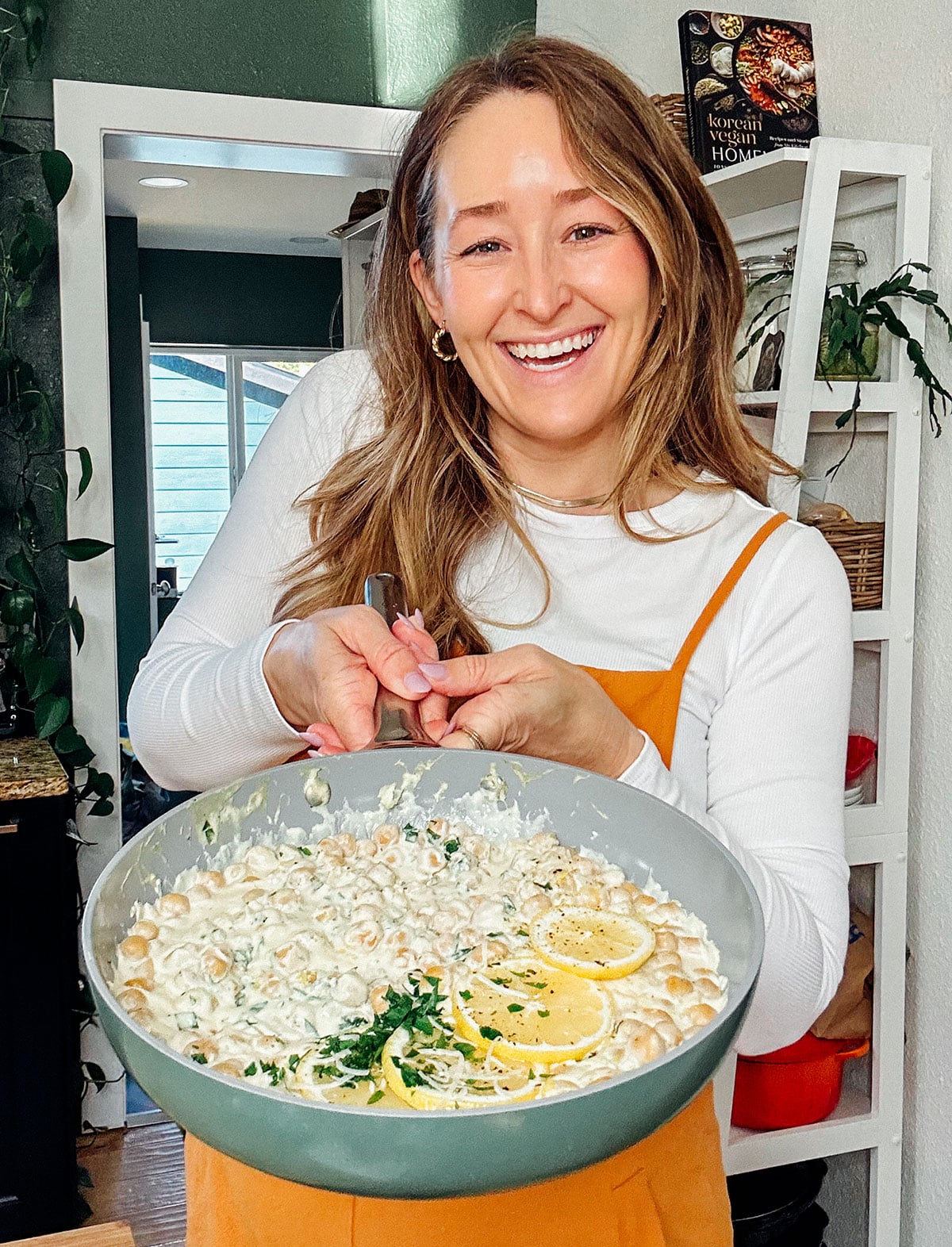 A woman in an orange apron smiles and holds a large bowl of high protein creamy pasta garnished with lemon slices, herbs, and chickpeas in a kitchen.