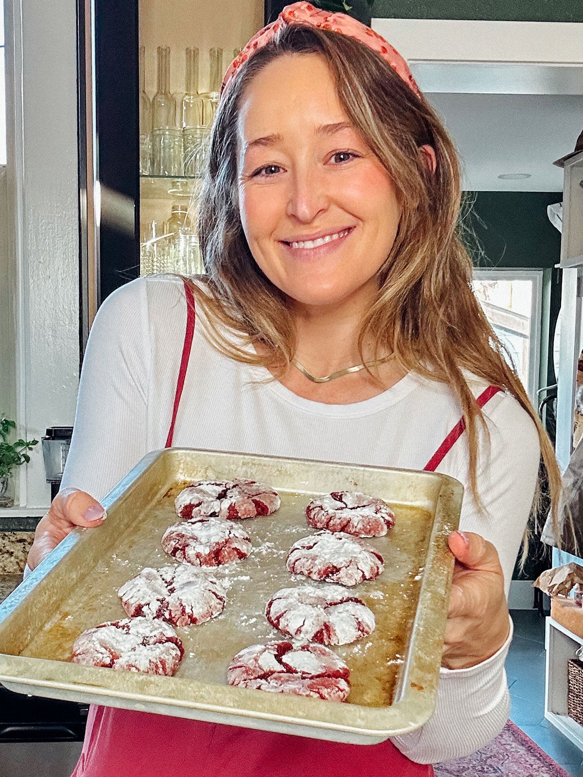 A woman in a white shirt and pink headband holds a tray of freshly baked vegan red velvet crinkle cookies dusted with powdered sugar.