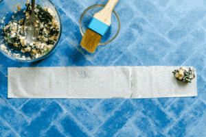 A bowl of spinach and cheese filling, a pastry brush, and a strip of dough with filling at one end are on a blue tiled surface.