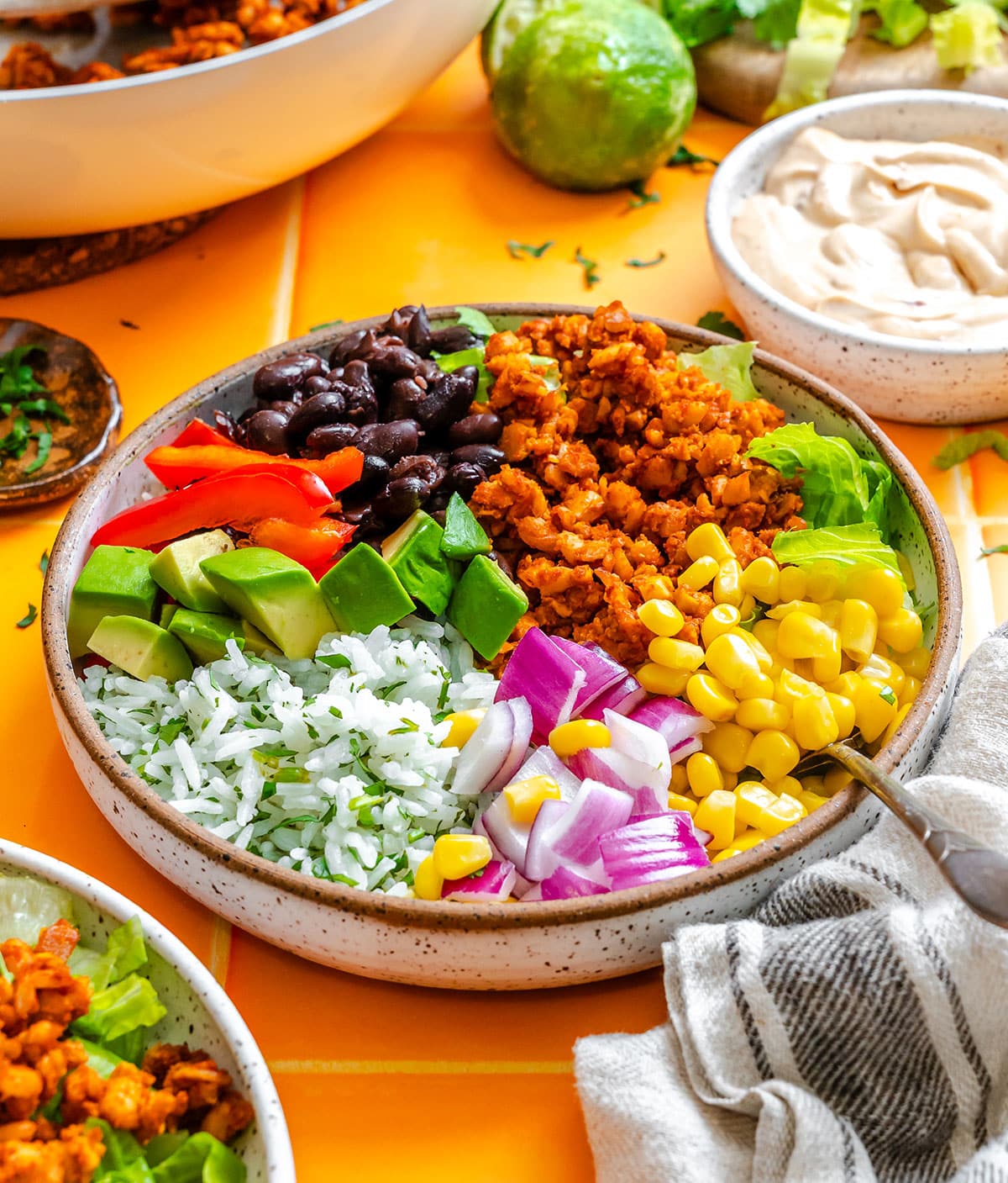A bowl filled with rice, black beans, corn, diced avocado, chopped red onion, red bell pepper, seasoned ground protein, and leafy greens on a yellow table.