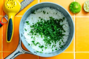 A saucepan containing cooked white rice topped with chopped fresh herbs, placed on an orange tiled surface with a lime, zester, and juicer nearby.