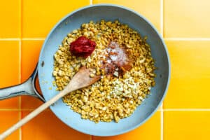 A frying pan with cooked lentils, chopped garlic, tomato paste, spices, and a wooden spoon on a bright orange tiled surface.