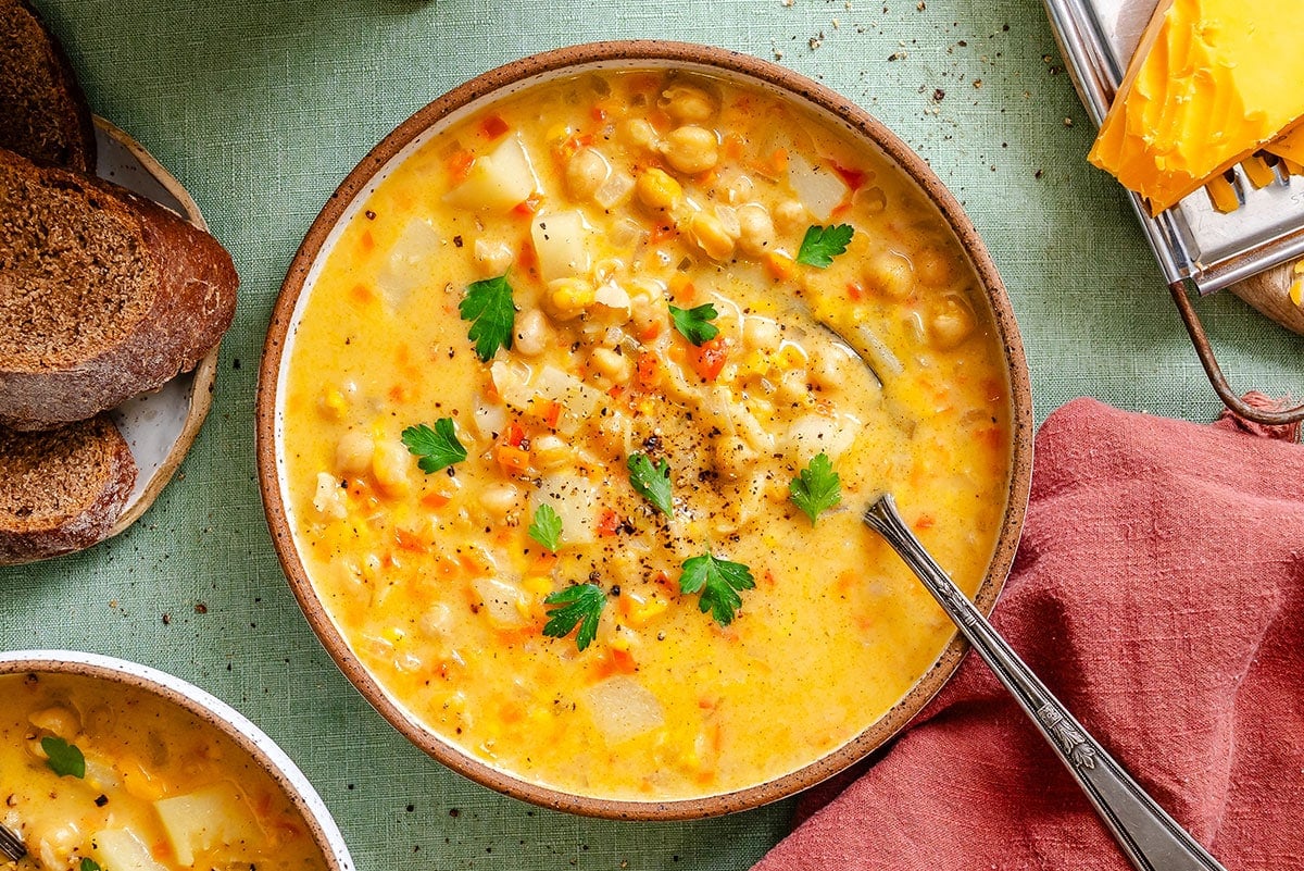 A bowl of chickpea and vegetable soup garnished with parsley, with a spoon inside, served next to slices of bread and a napkin on a green tablecloth.