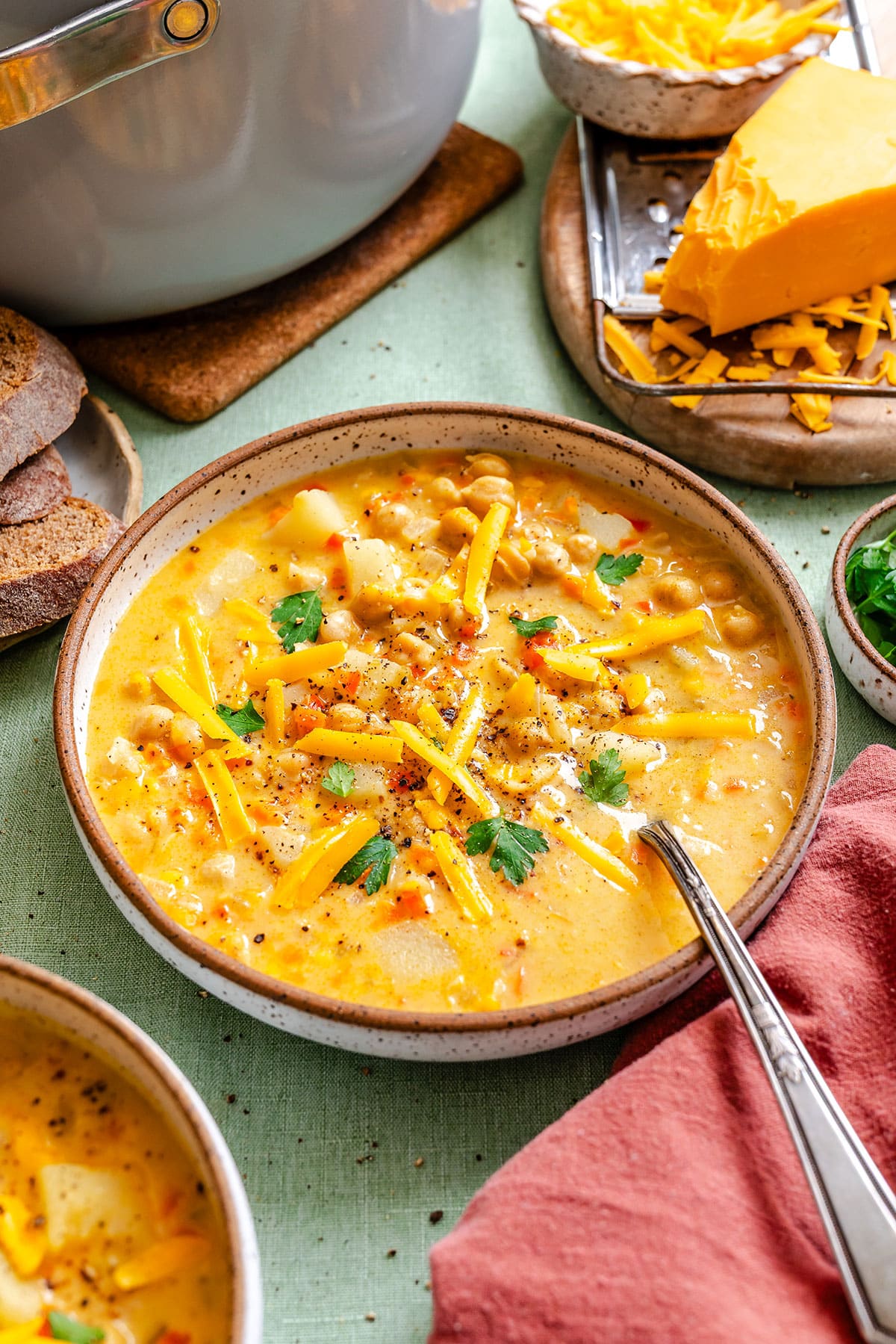 A bowl of cheesy chickpea chowder soup garnished with shredded cheese and parsley, with a spoon, bread slices, and a block of cheese in the background.