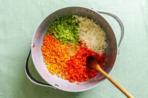 A pot containing chopped celery, onions, carrots, and red bell pepper arranged in sections, with a wooden spoon resting inside.