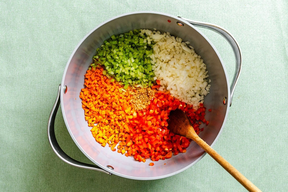 A pot containing chopped celery, onions, carrots, and red bell pepper arranged in sections, with a wooden spoon resting inside.