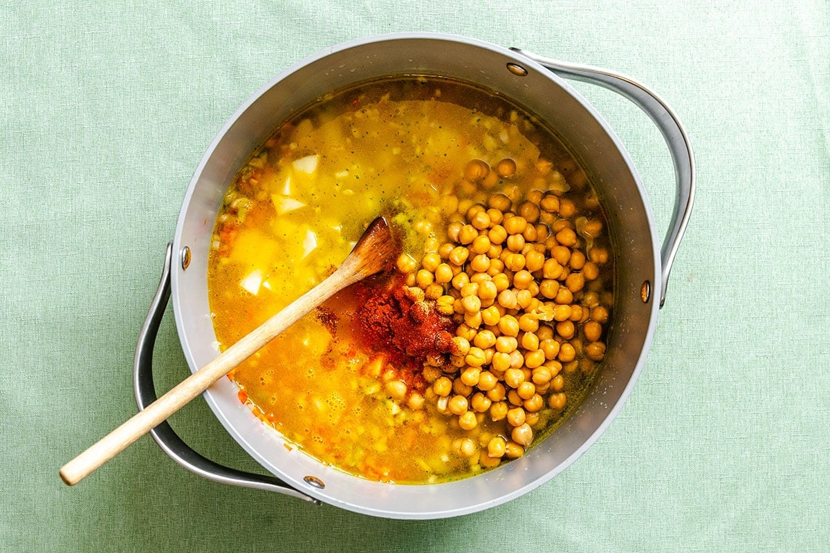A pot with a wooden spoon contains chickpeas, diced potatoes, broth, and spices on a light green surface.