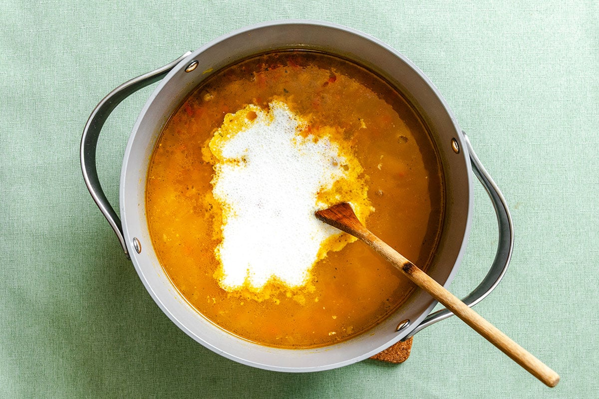 A large pot of chickpea chowder soup with a splash of cream being stirred by a wooden spoon, set on a light green surface.