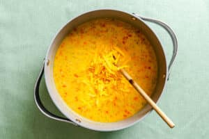 A large pot of creamy chickpea chowder soup with chopped vegetables and shredded cheddar cheese on top, being stirred with a wooden spoon.