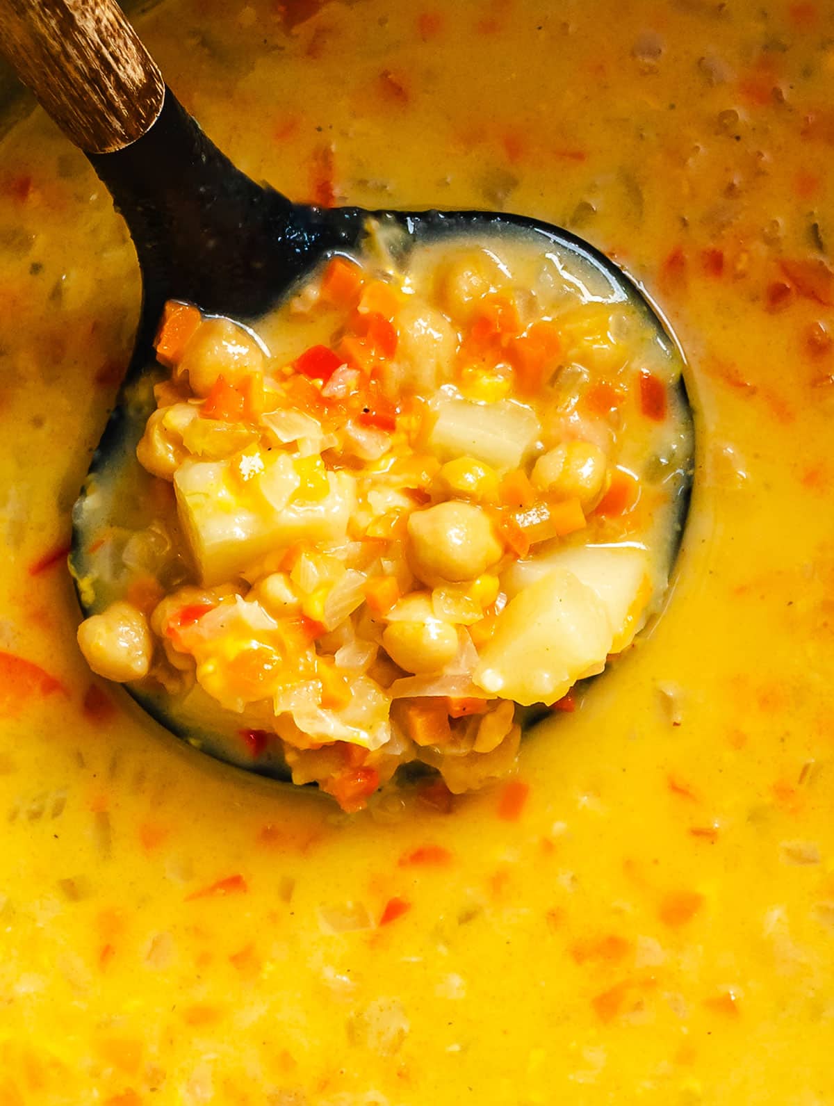 A close-up of a ladle filled with a thick chickpea chowder soup containing chickpeas, potatoes, carrots, and other diced vegetables.
