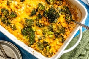 A baked casserole dish filled with broccoli, chickpeas, rice, and melted cheddar cheese, with a serving spoon on the side.