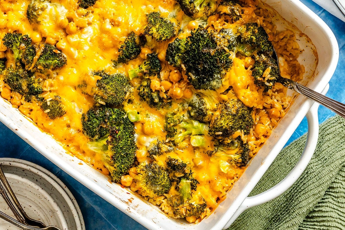 A baked casserole dish filled with broccoli, chickpeas, rice, and melted cheddar cheese, with a serving spoon on the side.