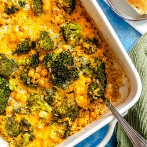 A baked casserole with broccoli, chickpeas, rice, and melted cheddar cheese in a white dish, with a serving spoon and bread slices on the side.