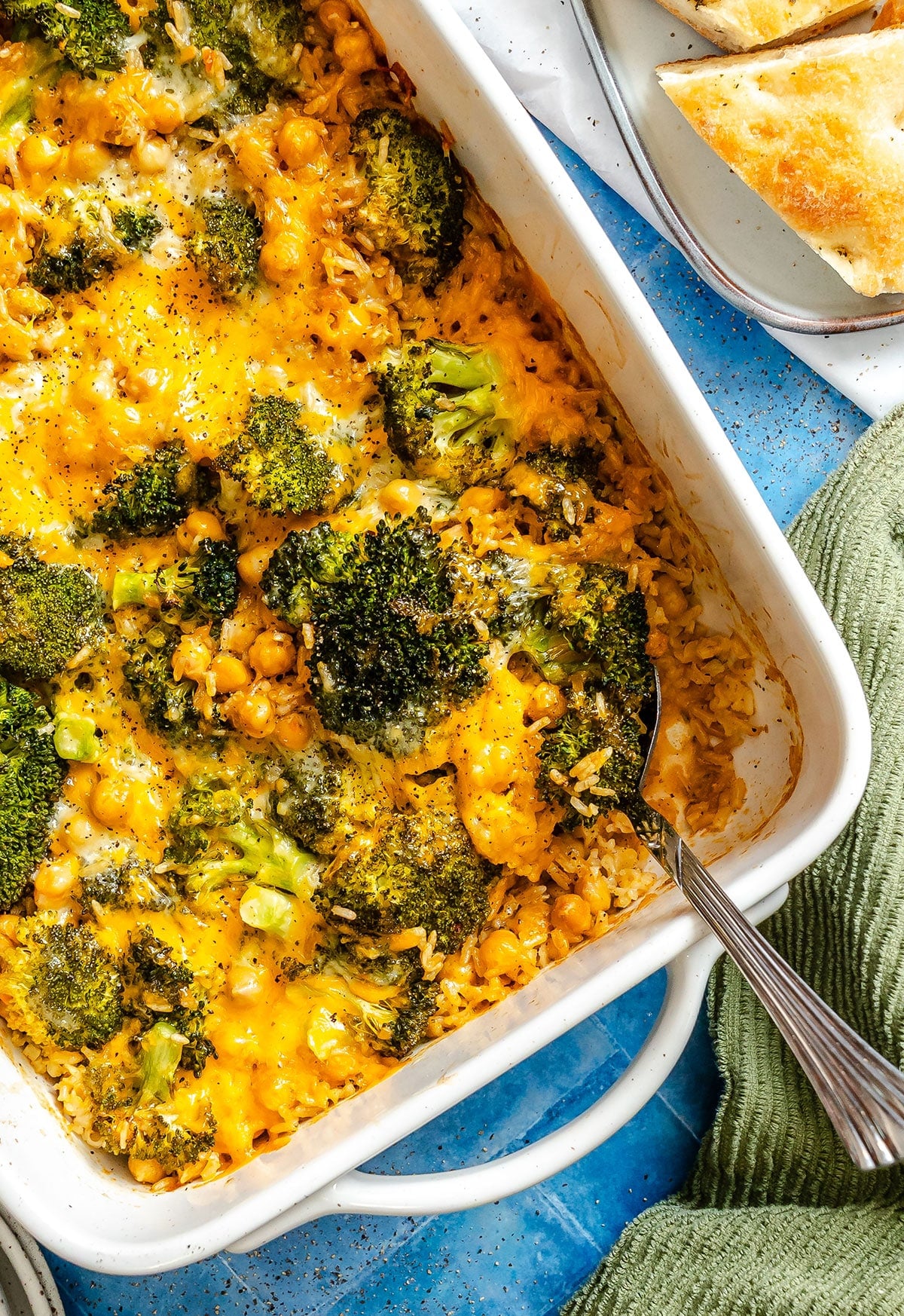 A baked casserole with broccoli, chickpeas, rice, and melted cheddar cheese in a white dish, with a serving spoon and bread slices on the side.