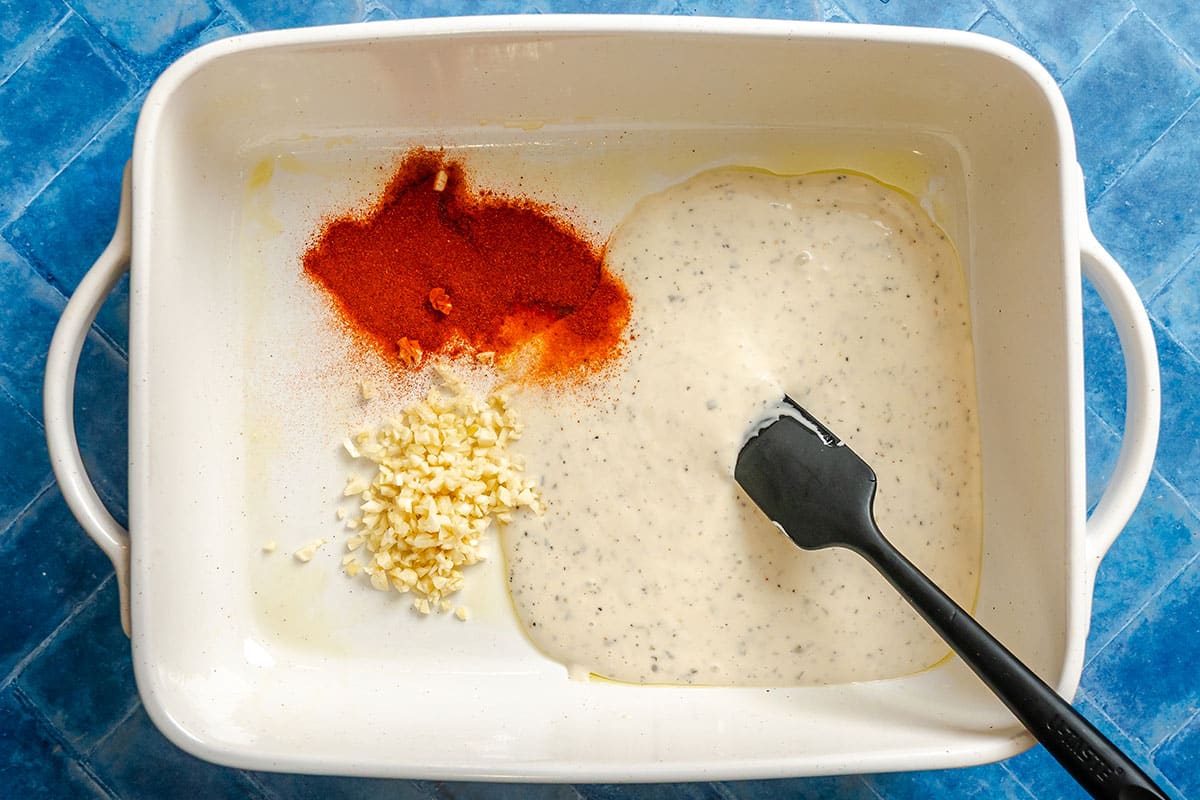 A white baking dish with chopped garlic, paprika, and creamy sauce being mixed with a black spatula, on a blue tiled surface.