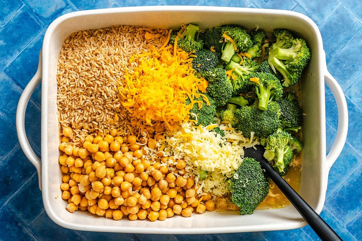 Rectangular white dish containing uncooked brown rice, shredded cheddar cheese, broccoli florets, chickpeas, and shredded mozzarella cheese on a blue tile surface.