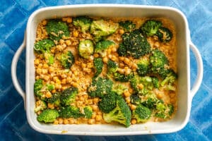 A baking dish filled with cooked broccoli florets, chickpeas, and a creamy sauce, set on a blue tiled surface.