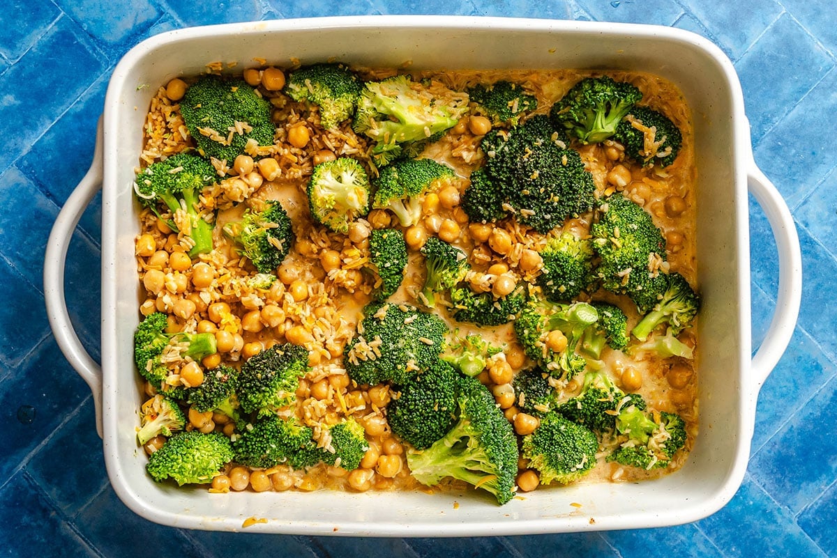 A baking dish filled with cooked broccoli florets, chickpeas, and a creamy sauce, set on a blue tiled surface.