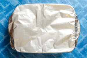 A rectangular baking dish covered with aluminum foil sits on a blue tiled surface.